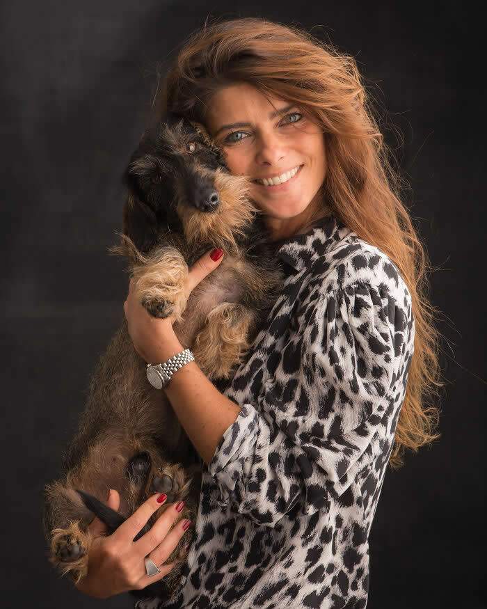 A smiling woman gently holds a small wire-haired dog close to her chest, both facing the camera, captured in soft studio lighting against a dark background, expressing warmth, comfort, and affectionate companionship.