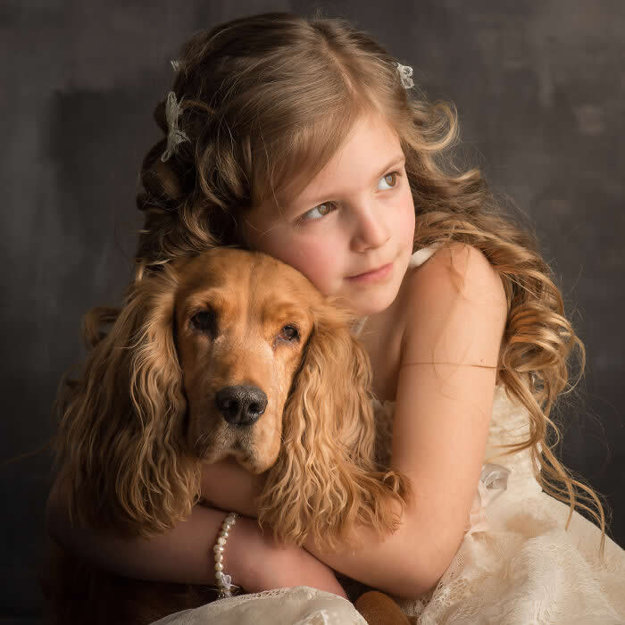 A young girl softly embraces a golden cocker spaniel, resting her cheek against the dog’s head, captured in warm studio lighting, expressing innocence, tenderness, and a deep emotional bond between child and animal.