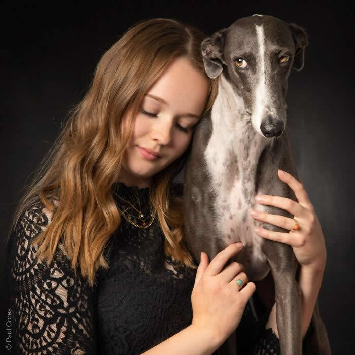 A young woman softly leans her head against a slender grey dog while holding it close, captured in warm studio lighting against a dark background, expressing calm affection, trust, and a quiet human–animal bond.