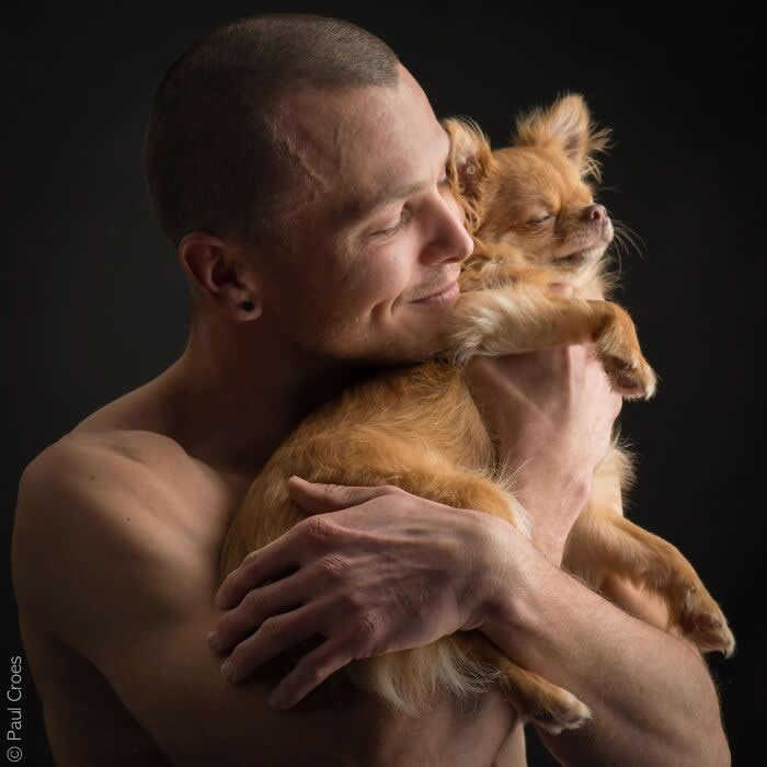 A man gently embraces a small dog close to his chest, both with eyes closed, captured in soft studio lighting that highlights warmth, trust, and a deep emotional connection between human and animal.