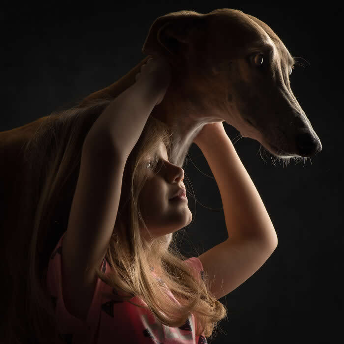 A young girl gently lifts a slender dog above her head, both softly lit against a dark background, creating a tender silhouette that conveys protection, trust, and a deep emotional connection between child and animal.