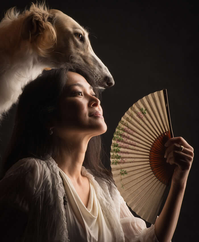 A softly lit portrait of a woman holding a decorative fan while a large dog rests its head gently above hers, both gazing upward, creating a serene composition that conveys elegance, trust, and quiet companionship.