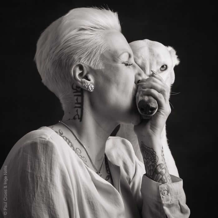 A black-and-white portrait of a woman with short hair gently kissing a white dog’s face while holding its muzzle, captured in soft studio lighting, expressing deep affection, trust, and an intimate human–animal bond.