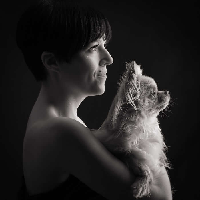 A black-and-white side-profile portrait of a woman holding a small fluffy dog, both gazing in the same direction, softly lit against a dark background, conveying unity, calm, and a shared emotional connection.