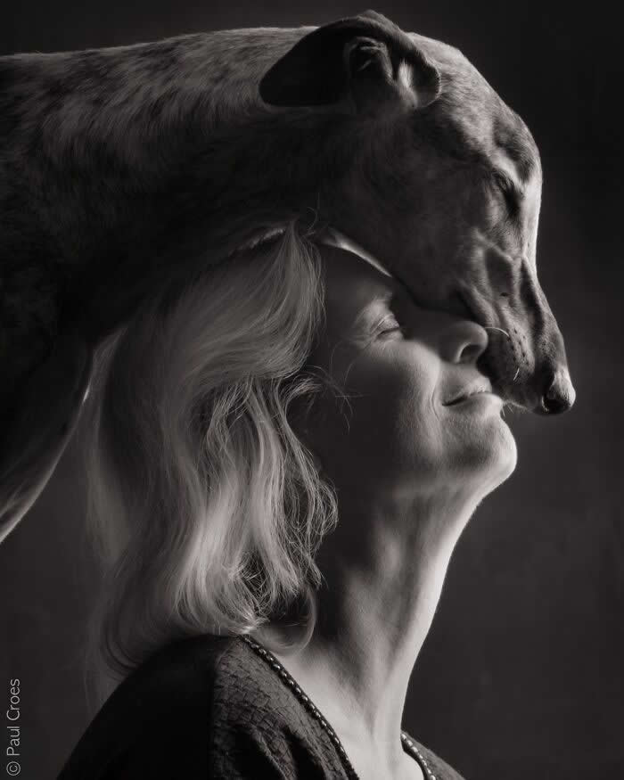A black-and-white portrait of a dog gently resting its head against a woman’s face, both with eyes closed, captured in soft studio lighting, expressing deep trust, affection, and a serene human–animal bond.