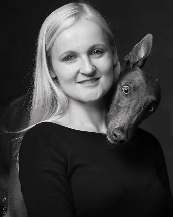 A black-and-white portrait of a smiling woman with a slender dog resting its head over her shoulder, both facing the camera, captured in soft studio lighting, expressing companionship, trust, and quiet connection.