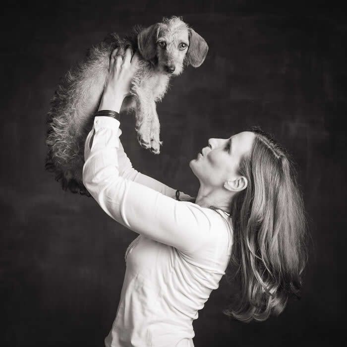 Black-and-white portrait of a woman holding a small dog up in the air, gazing at it with affection while the dog looks toward the camera, capturing a playful and tender human-animal connection.