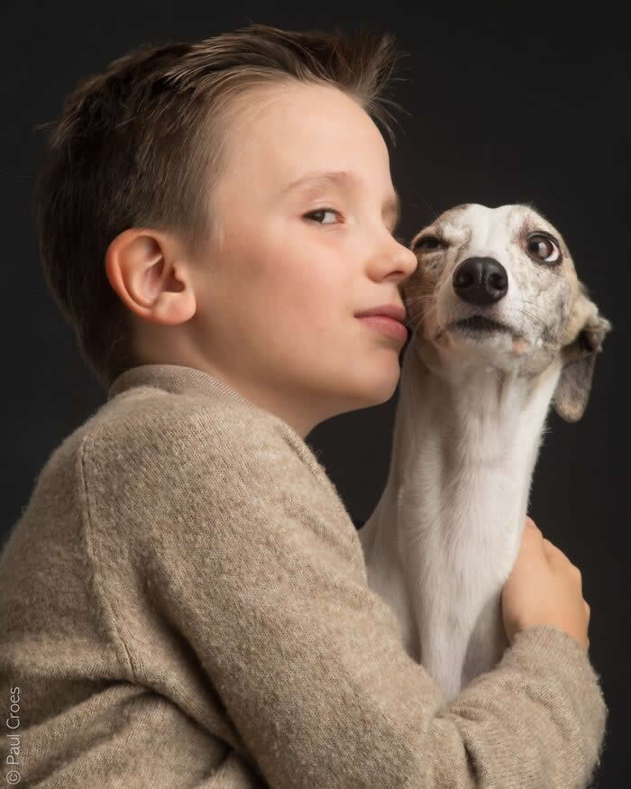 A young boy gently hugs a slender dog close to his face, both looking toward the camera, captured in soft studio lighting that highlights trust, closeness, and emotional connection.