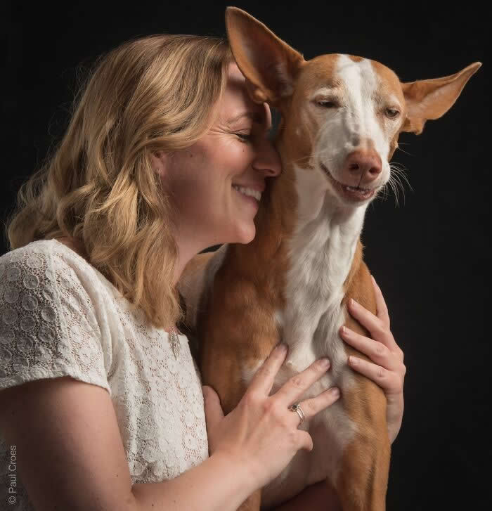 A smiling woman gently embraces a tall, slender dog, both with relaxed expressions, captured in soft studio lighting against a dark background, conveying warmth, joy, and a deep emotional connection.
