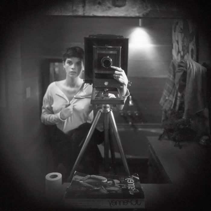 Black-and-white mirror self-portrait of photographer Sally Mann standing behind a large format camera on a tripod in a dim interior room, with a magazine and paper roll visible in the foreground.