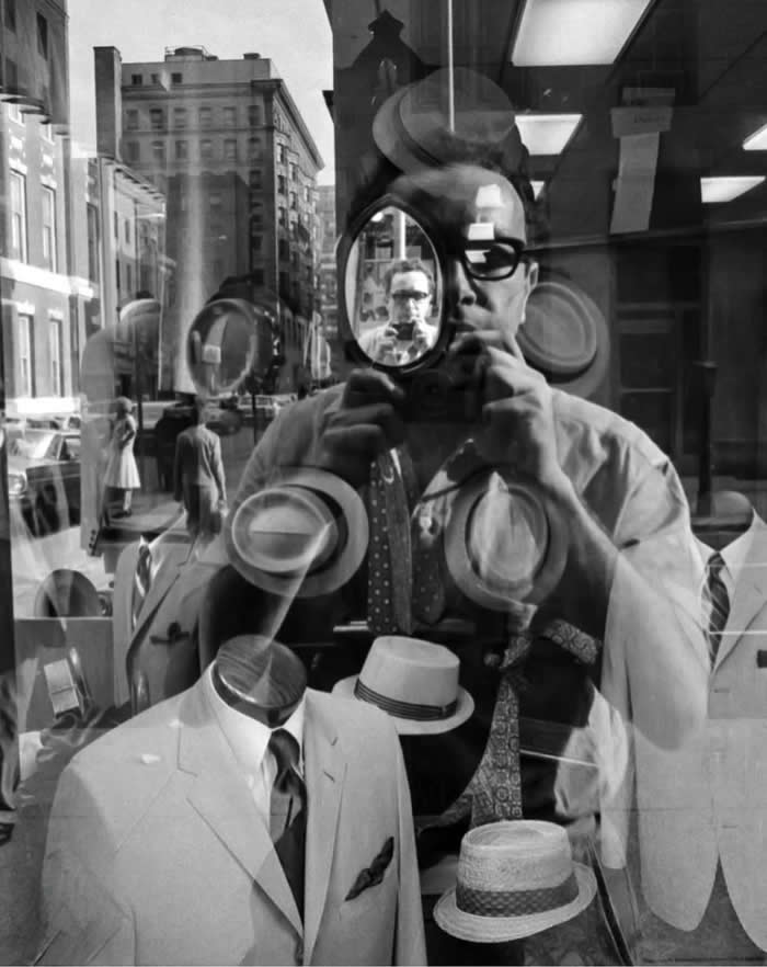 Black-and-white reflection self-portrait of photographer Harold Feinstein holding a camera in a storefront window, layered with mannequins, hats, ties, and city street reflections.