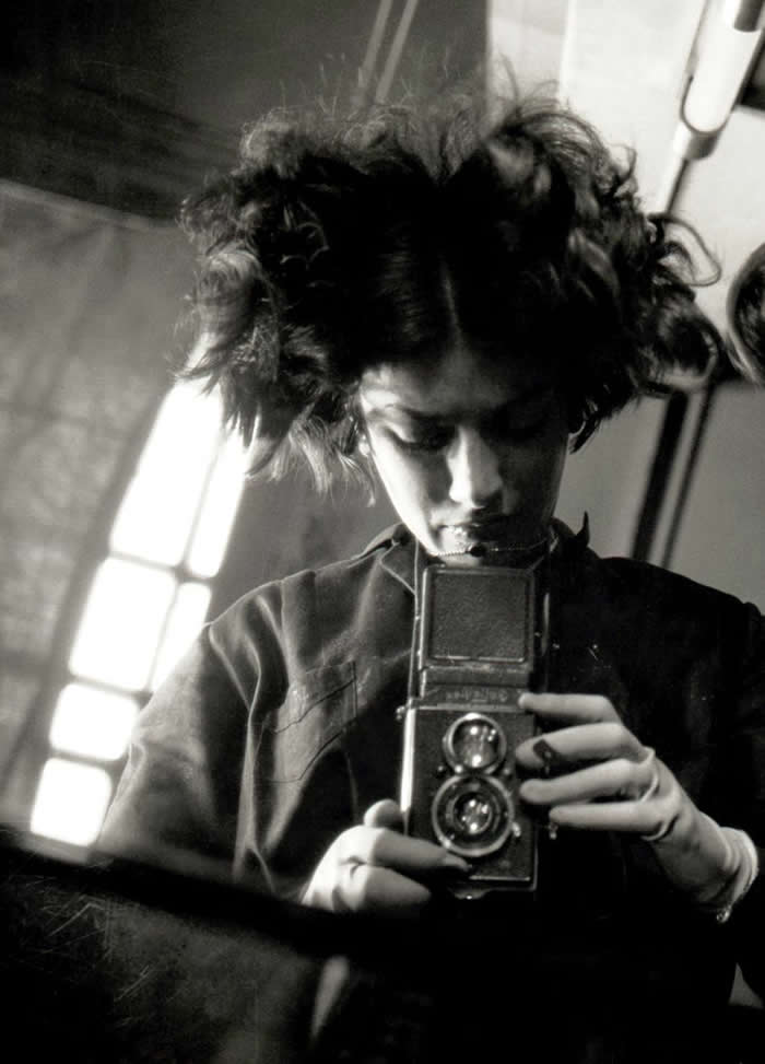 Black-and-white mirror self-portrait of photographer Eva Besnyö looking downward while holding a twin-lens reflex camera, with dramatic light from a nearby window illuminating her face and hair.