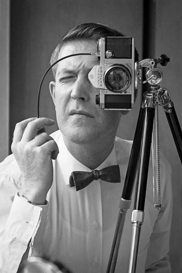 Black-and-white self-portrait of photographer Jack Sharp wearing a bow tie, squinting through a camera mounted on a tripod while holding a cable release in one hand.