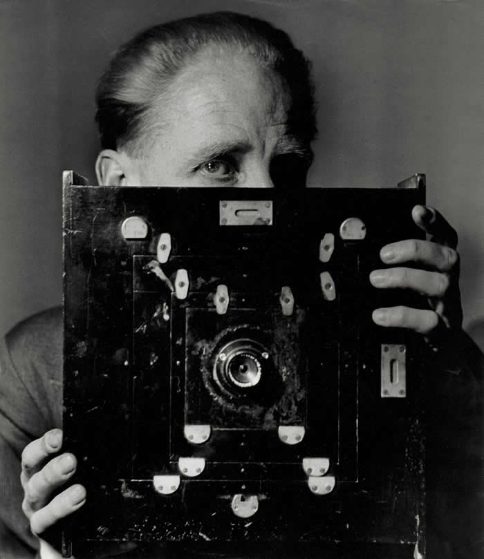 Black-and-white self-portrait of photographer Bill Brandt partially hidden behind a large vintage camera, with one eye visible above the camera body and hands gripping both sides.