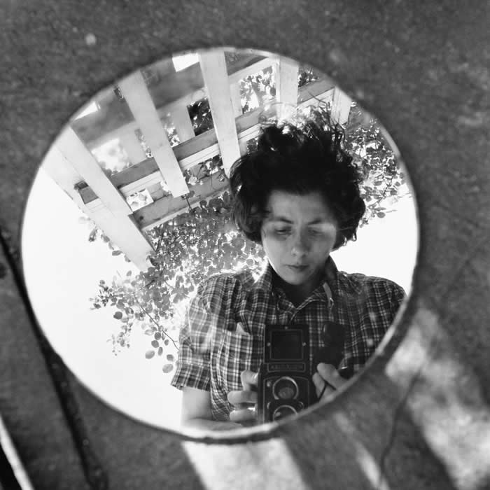 Black-and-white mirror self-portrait of photographer Vivian Maier holding a twin-lens reflex camera, reflected in a round mirror outdoors beneath wooden beams and leafy branches.