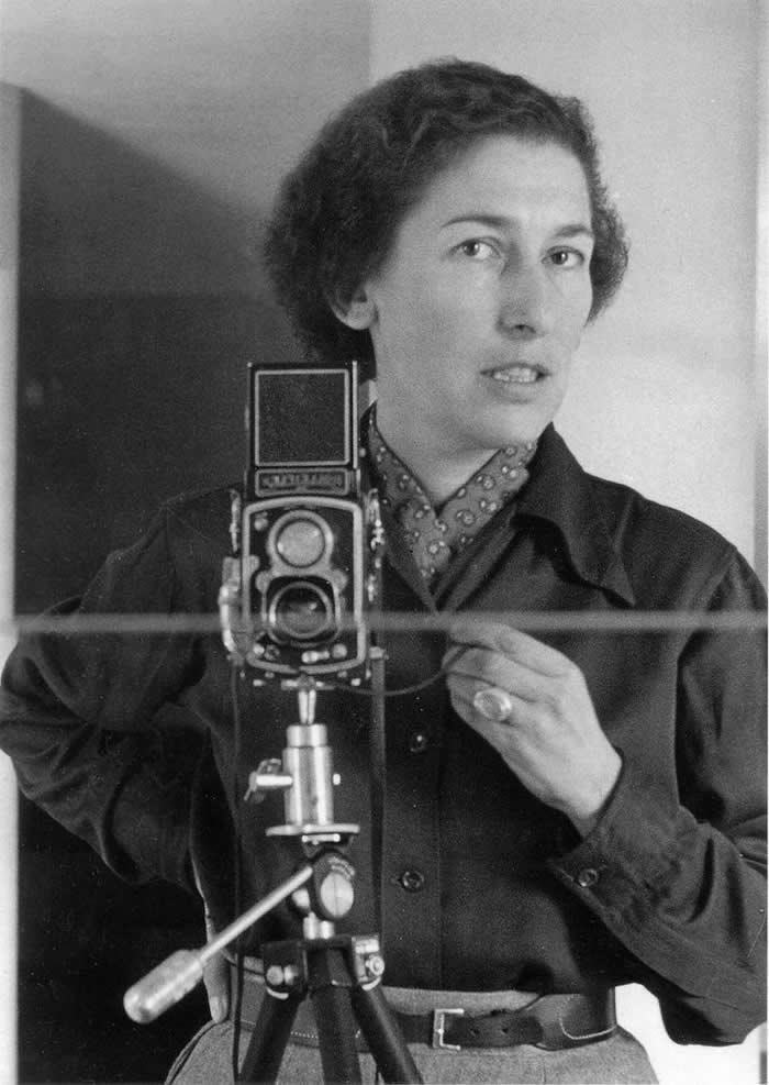 Black-and-white mirror self-portrait of photographer Gisèle Freund standing beside a twin-lens reflex camera on a tripod, wearing a dark jacket and patterned scarf while looking toward the mirror.