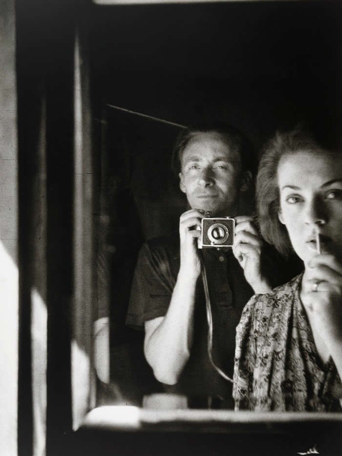 Black-and-white mirror self-portrait of photographer Albert Tucker holding a camera beside his wife, who stands in the foreground with a cigarette, both reflected in a dimly lit mirror.