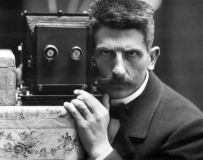 Black-and-white self-portrait of photographer Frédéric Boissonnas posing beside a vintage box camera, staring intensely toward the viewer, with one hand resting near the camera.