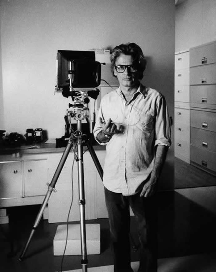 Black-and-white self-portrait of photographer Richard Avedon standing in a studio beside a large format camera on a tripod, wearing glasses and holding a remote shutter cable.