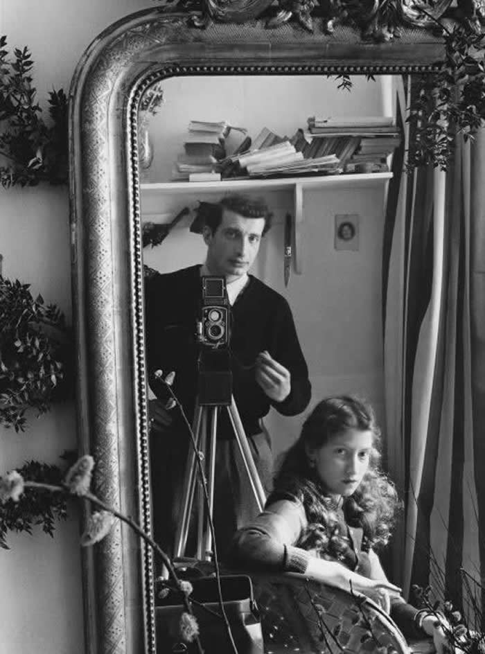 Black-and-white mirror self-portrait of photographer Édouard Boubat standing behind a camera on a tripod, with companion Lella seated in front, reflected in an ornate framed mirror.