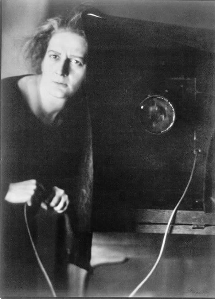 Black-and-white self-portrait of photographer Lotte Jacobi standing beside a large vintage camera, leaning forward with an intense expression while holding a shutter release cable.