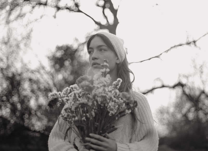 Black and white portrait of a young woman wearing a knit hat and holding a bouquet of flowers, standing outdoors with soft light and blurred trees creating a calm, poetic atmosphere.