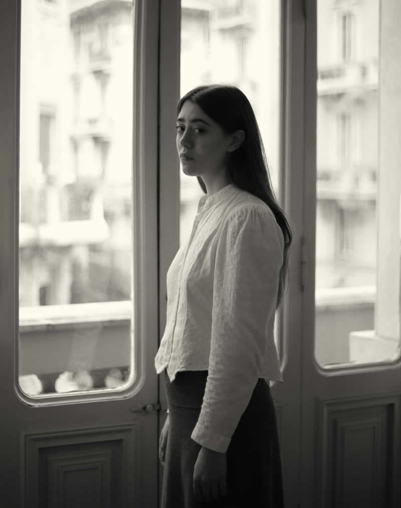 Soft black and white portrait of a woman standing by a window, looking back over her shoulder, with natural light and blurred city architecture in the background creating a quiet, reflective mood.