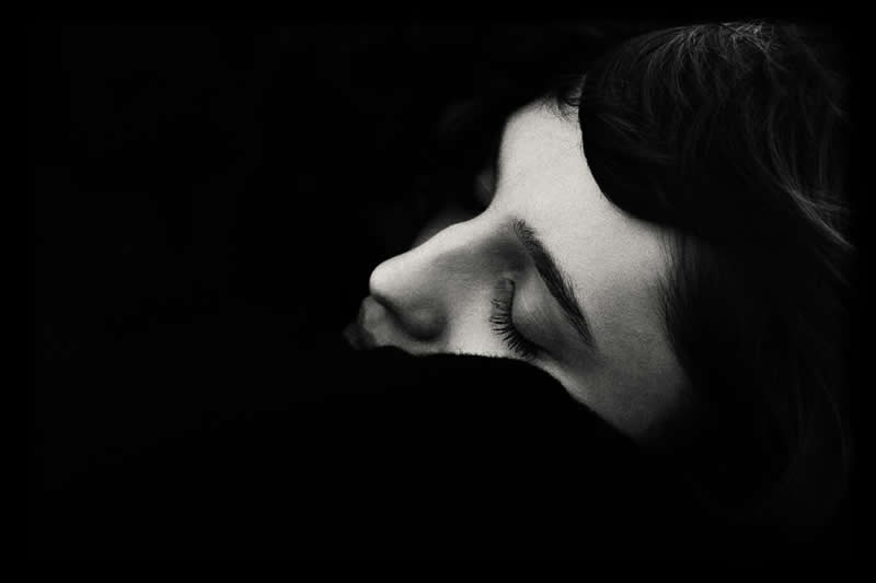 High-contrast black and white close-up of a person’s face in profile with eyes closed, partially hidden in shadow, creating a quiet and introspective mood.