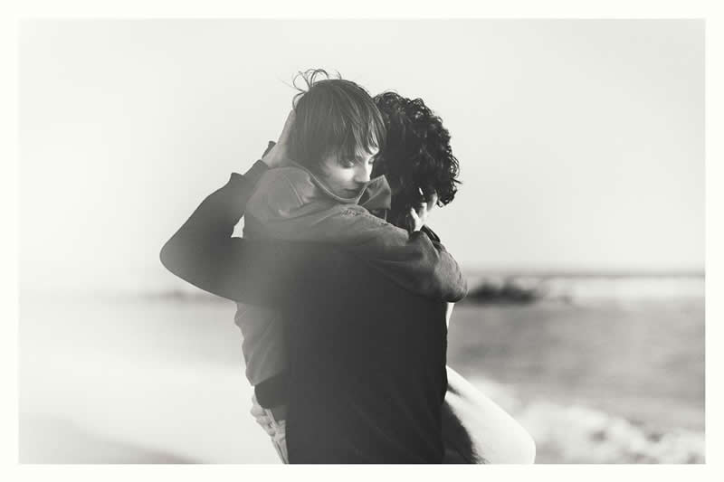 Black and white photo of two people embracing by the seaside, with soft haze and gentle light creating an emotional and intimate atmosphere.
