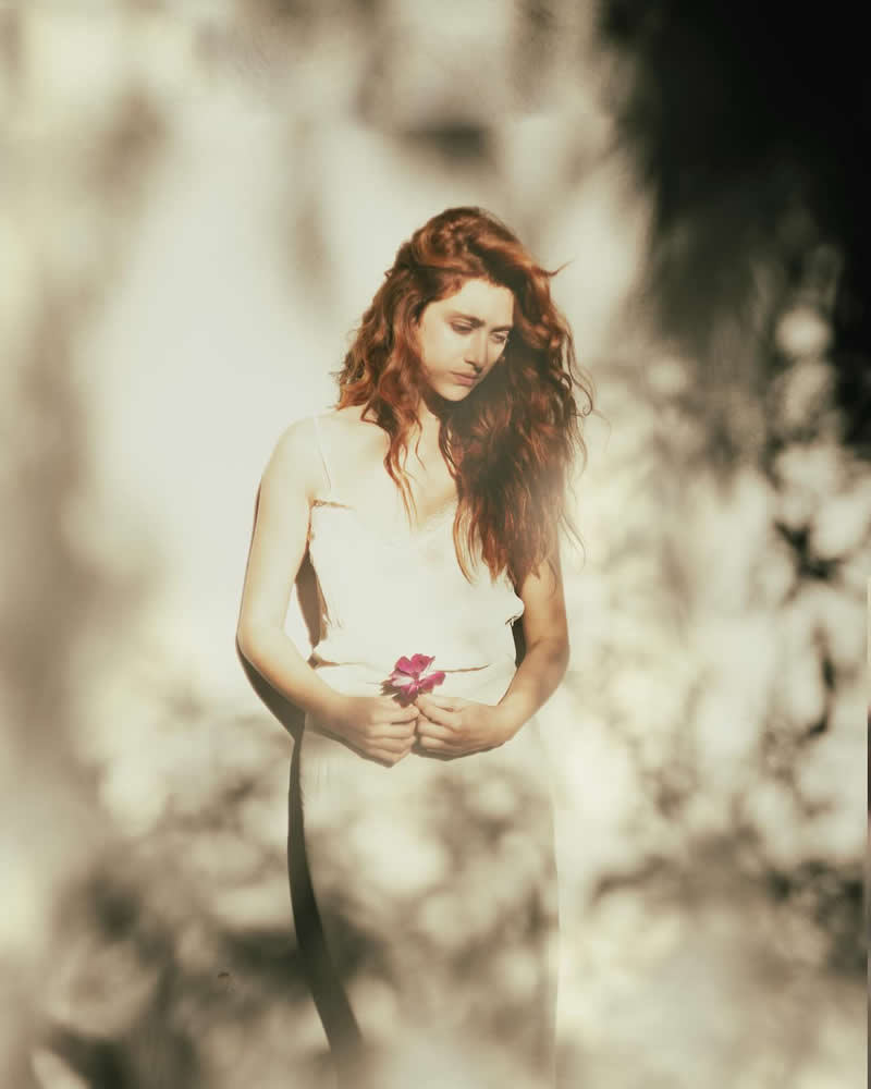 Soft, warm-toned portrait of a woman standing outdoors in sunlight, gently holding a small flower, surrounded by blurred foliage and a dreamy, ethereal atmosphere.