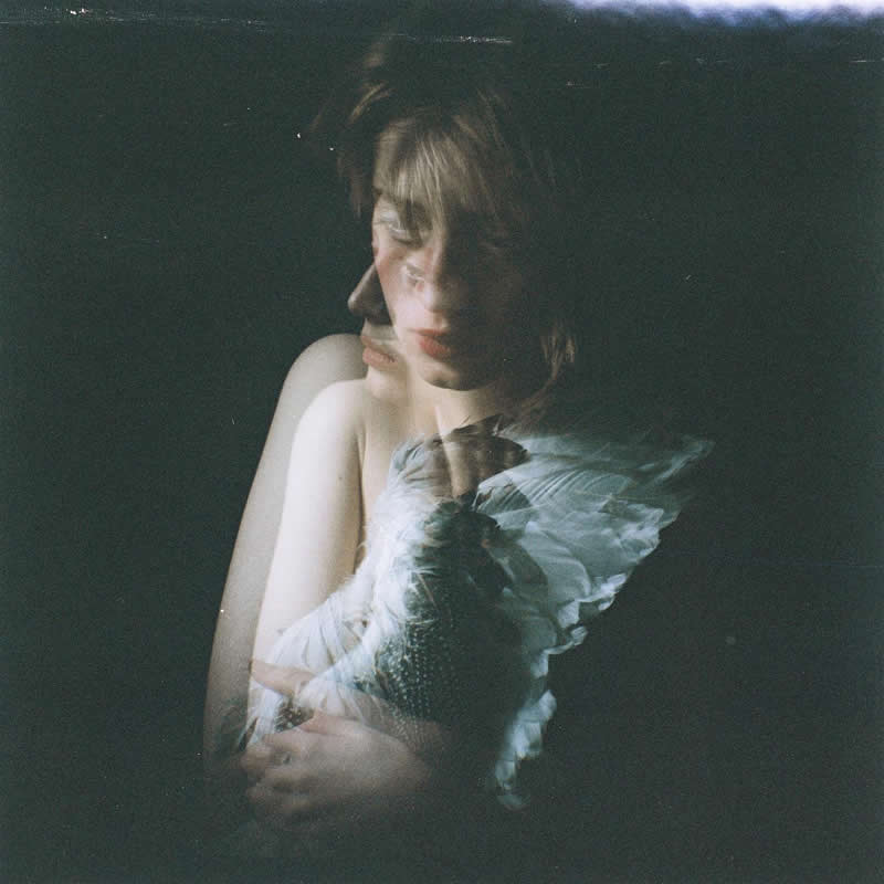 Dark, moody double exposure portrait of a woman with overlapping profiles and a soft feather detail, creating a surreal and introspective atmosphere with low light and muted tones.