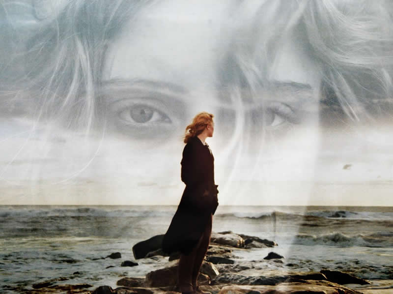 Double exposure image of a woman standing on a rocky shoreline facing the sea, overlaid with a large, faded portrait of eyes, creating a surreal and emotional coastal scene.