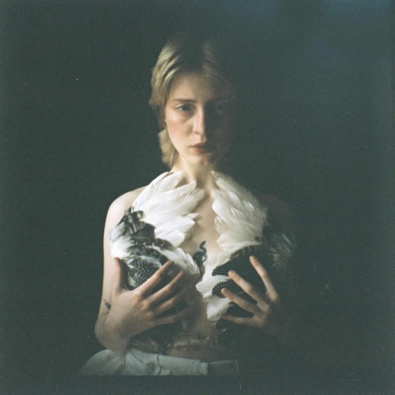 Moody, low-light portrait of a person holding white feathers close to their chest, softly illuminated against a dark background, creating a delicate and symbolic atmosphere.