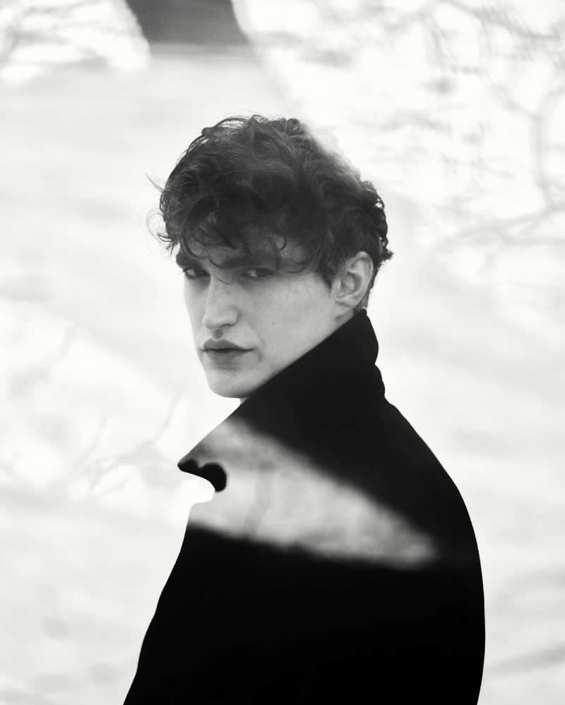 Black and white portrait of a person in a dark coat looking back over their shoulder, with soft background blur and a subtle overlay creating a moody, reflective atmosphere.