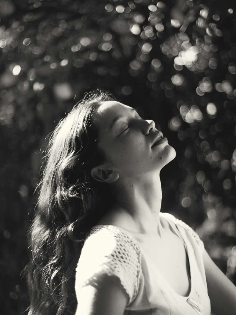 Black and white portrait of a woman with her head tilted back and eyes closed, bathed in sunlight with soft bokeh in the background, creating a serene and uplifting mood.
