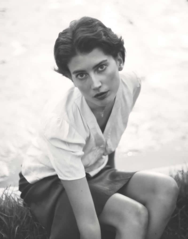 Black and white portrait of a woman sitting on the ground, looking directly up at the camera with an intense gaze, surrounded by soft, minimal background tones.