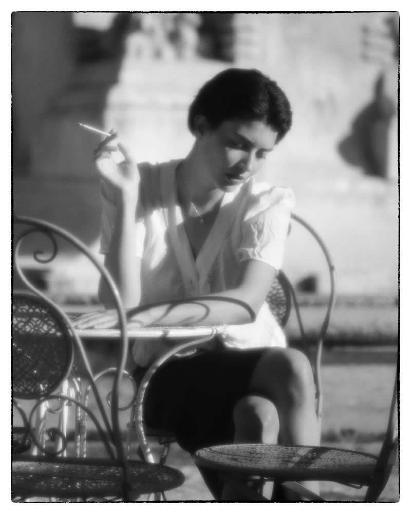 Black and white vintage-style portrait of a woman sitting at an outdoor café table, holding a cigarette, captured in soft light with a dreamy, nostalgic atmosphere.
