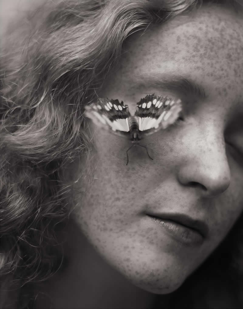 Black and white close-up portrait of a freckled person with closed eyes, featuring a butterfly resting on their eyelid, creating a surreal and poetic mood.