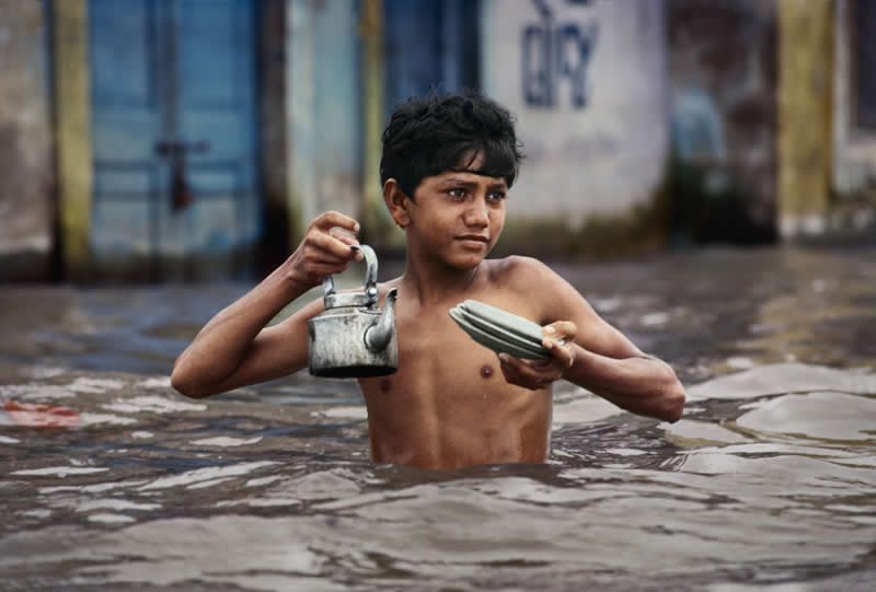 Tea Vendor carries tea kettle in waist deep monsson water, Porbandar, India by Steve McCurry - Iconic Color Photos by Legendary Photographers
