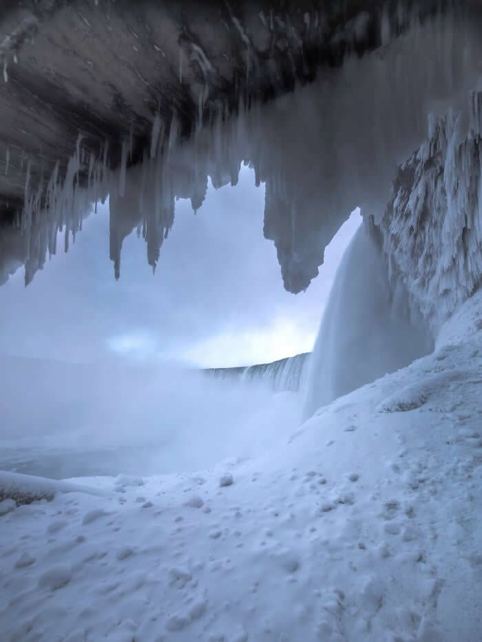 Icicles hang from a frozen cave above Niagara Falls, framing a snowy landscape and misty waterfall cascading through icy winter conditions.
