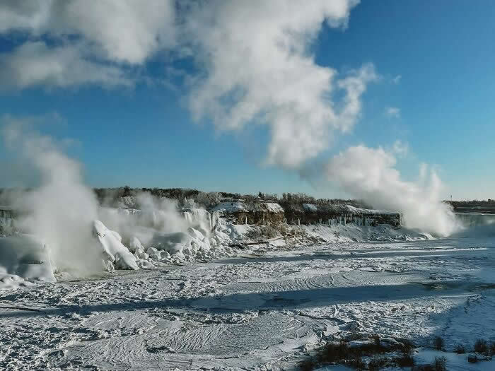 Steam-like mist rises above frozen Niagara Falls and icy riverbanks under a bright blue sky, creating a dramatic winter landscape filled with snow and frozen textures.