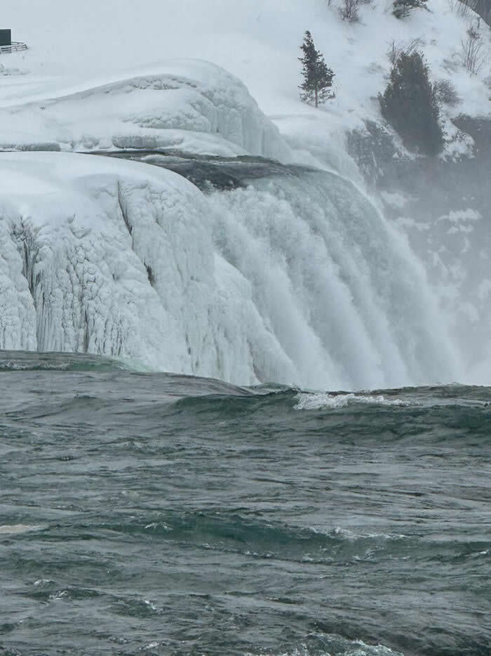 Close-up view of icy Niagara Falls cascading over snow-covered cliffs, with dark rushing water contrasting against frozen winter formations and misty surroundings.