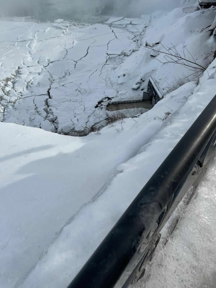 Snow-covered walkways and icy cliffs overlook cracked frozen water near Niagara Falls, creating a stark and dramatic winter scene from a high viewpoint.