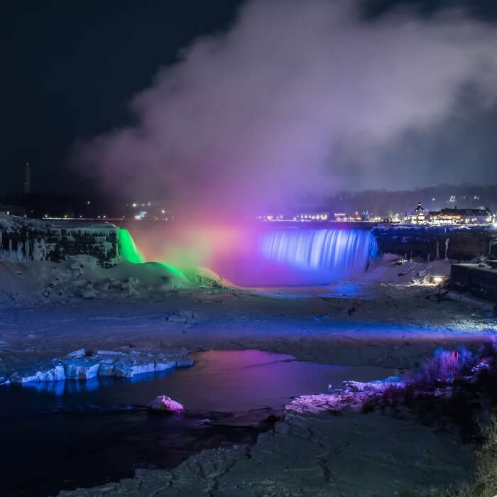 Niagara Falls glows with colorful green, purple, and blue lights at night, surrounded by icy waters, frozen shoreline, and rising mist under a dark winter sky.