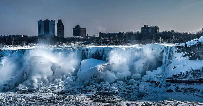Icy Niagara Falls crashes through massive snow-covered formations with mist rising into the cold air, while city buildings stand beneath a deep winter sky.