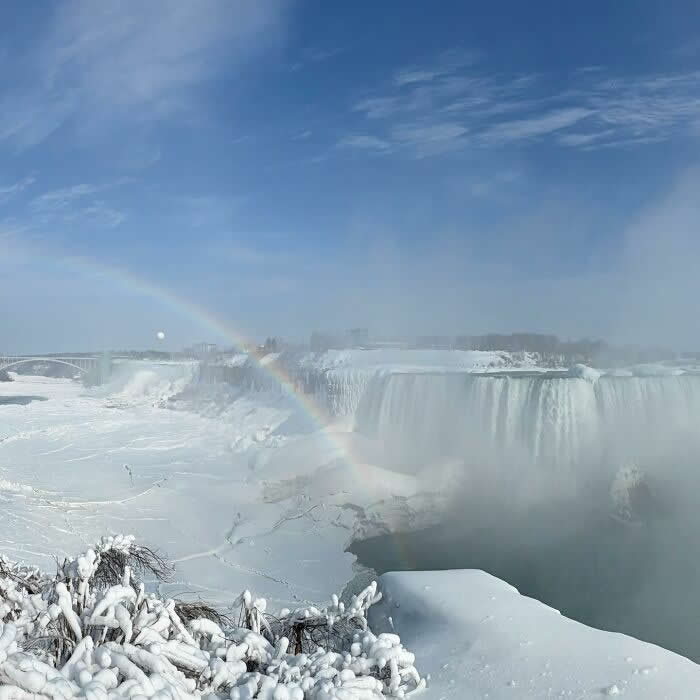 A faint rainbow appears above snowy Niagara Falls as icy cliffs, frozen trees, and drifting mist glow beneath a bright blue winter sky.