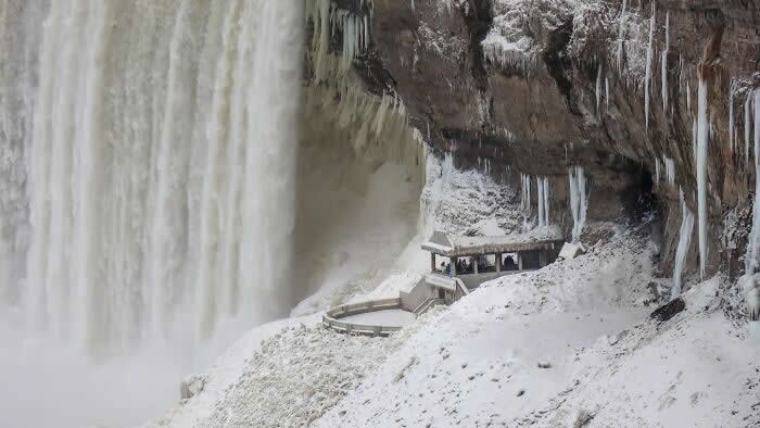A snowy viewing platform sits beneath towering Niagara Falls and icy cliffside formations, surrounded by frozen mist and hanging icicles in winter.