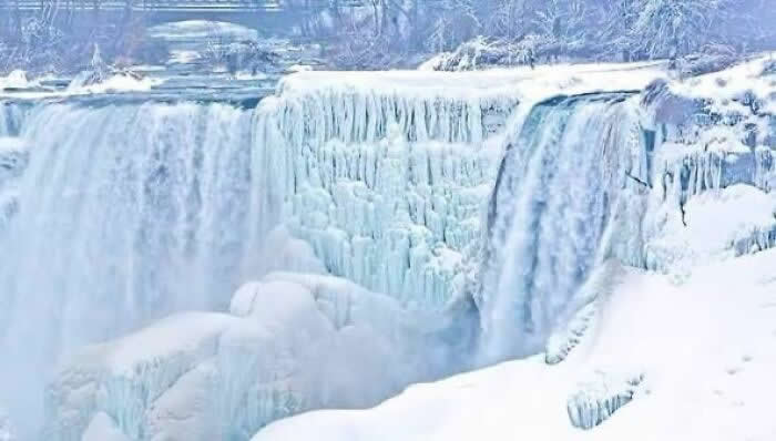 Frozen Niagara Falls pours over thick ice-covered cliffs and snow-covered formations, creating a bright winter scene filled with icy textures and cascading water.