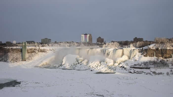 A wide winter view of Niagara Falls shows frozen waterfalls, snow-covered riverbanks, drifting mist, and distant city buildings beneath a calm gray sky.