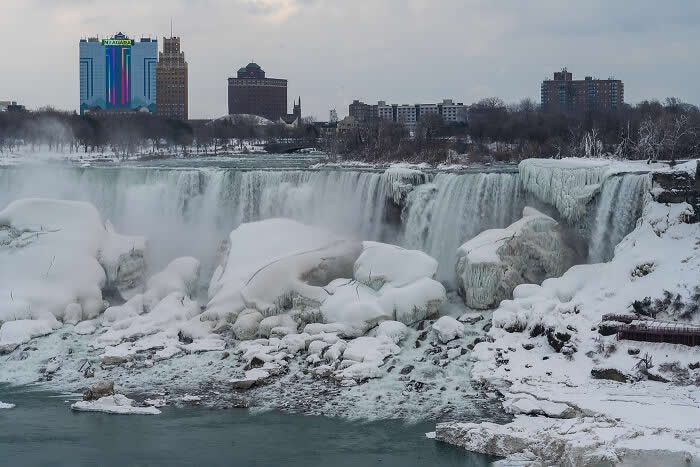 Snow-covered Niagara Falls cascades through icy rock formations with mist rising into a gray winter sky, while city buildings stand in the background.